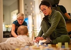 Eine junge Mutter sitzt auf dem Fußboden und spielt mit ihrem Baby mit Bauklötzen. Daneben kniet dem Baby zugewandt eine Familien-Gesundheits- und Kinderkrankenpflegerin.