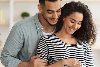Mann steht hinter seiner schwangeren Frau, umarmt sie und schaut auf den Babybauch. Die Frau umfasst den runden Bauch.