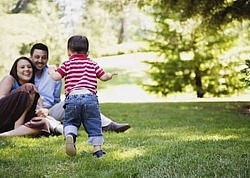 Eltern sitzen auf einer Wiese im Park, ihr Kleinkind läuft auf sie zu