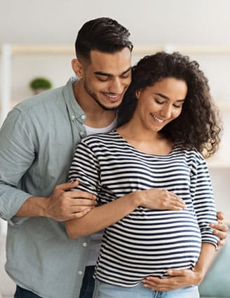 Mann steht hinter seiner schwangeren Frau, umarmt sie und schaut auf den Babybauch. Die Frau umfasst den runden Bauch.