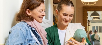 Junge Mutter begrüßt Besuchende der Frühen Hilfen an der Haustür und betrachtet mit ihr neutral das Baby