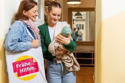 Junge Mutter begrüßt Besuchende der Frühen Hilfen an der Haustür und betrachtet mit ihr neutral das Baby
