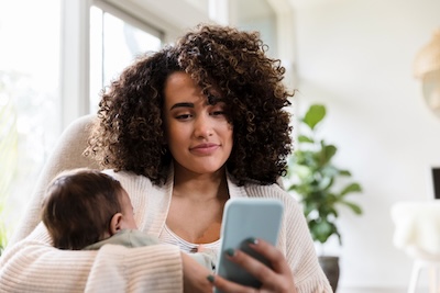 Eine Mutter hält ihr Baby auf dem Arm und in der anderen Hand ein Smartphone, auf das sie schaut.