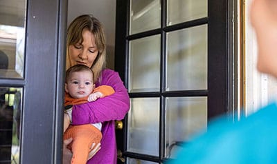 Eine Mutter öffnet einer Besucherin die Tür, sie hält ihr Neugeborenes auf dem Arm