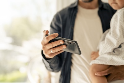 Vater trägt Baby auf dem Arm und hält in der anderen Hand ein Handy, auf das er schaut.