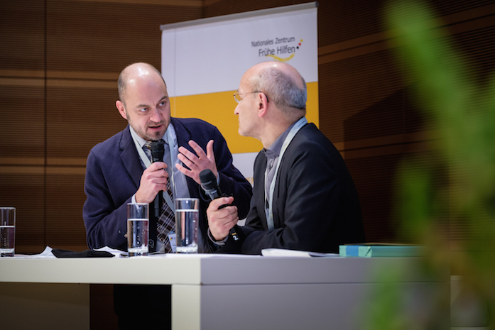 Prof. Dr. Michael Kölch und Prof. Dr. Albert Lenz stehen an einem Tisch auf der Bühne und diskutieren, dahinter ein Plakataufsteller