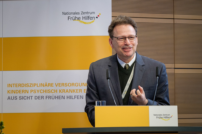 Prof. Dr. Daniel S. Schechter bei seinem Vortrag am Rednerpult, dahinter ein Plakataufsteller mit Logo des NZFH