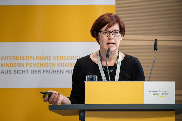 Prof. Dr. Sabine Wagenblass bei ihrem Vortrag am Rednerpult, dahinter ein Plakataufsteller mit Logo des NZFH