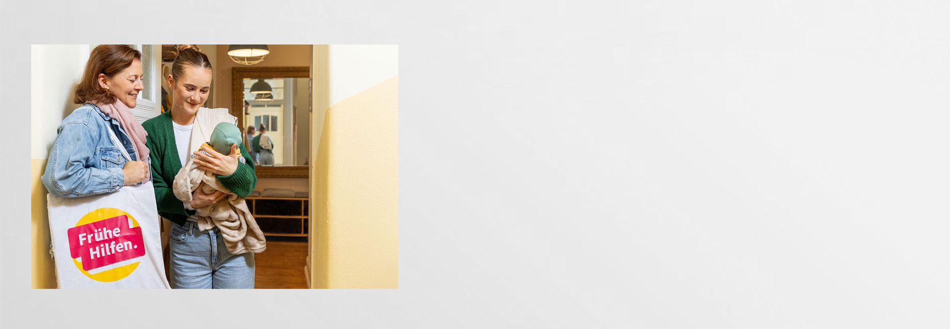 Junge Mutter begrüßt Besuchende der Frühen Hilfen an der Haustür und betrachtet mit ihr neutral das Baby