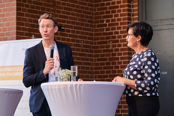 Peter Rölle-Dahl am Mikrofon an einem Stehtisch auf dem Podium mit Moderatorin Christiane Poertgen