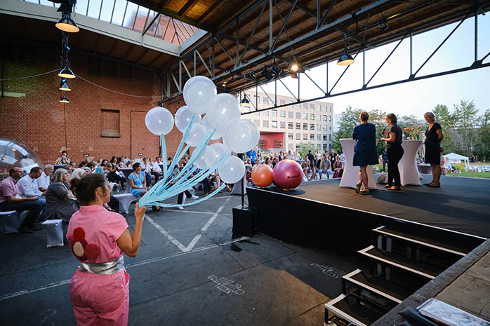 Mitglied von Kompanie Kopfstand e.V. mit Ballons an Bändern in der Hand neben dem Podium mit sitzenden Teilnehmenden im Hintergrund