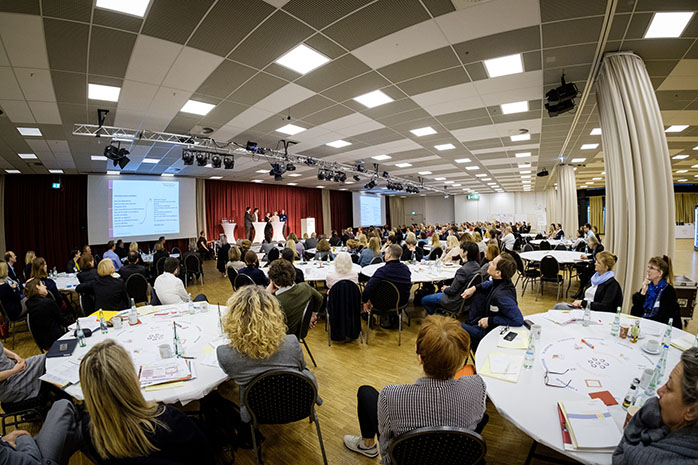 Teilnehmende sitzen in einem großen Raum an runden Tischen und Blicken zu den Referierenden, die auf einer Bühne stehen, rechts und links eine Präsentationswand