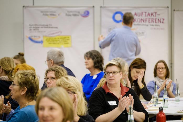 Teilnehmende sitzen an runden Tischen, manche klatschen, dahinter hält ein Konferenzmaler die Ergebnisse grafisch auf einem Plakat fest.