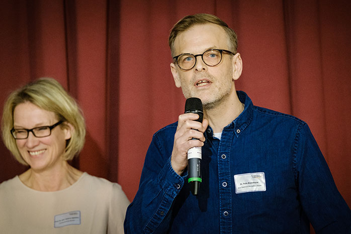 Dr. Felix Brandhorst steht vor rotem Vorhang und spricht in ein Mikrofon.