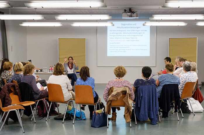 Sitzende Teilnehmende von hinten schauen nach vorne auf Prof. Dr. Leonie Herwartz-Emden die vor einer Pinnwand sitzt, neben ihr eine Projektion mit Texten an der Wand