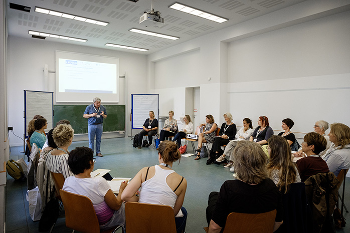 Prof. Dr. Patrick Meurs steht vor Teilnehmenden die in einem Halbkreis auf Stühlen sitzen, hinter ihm eine Tafel, Projektionswand sowie Pinnwände rechts und links neben ihm