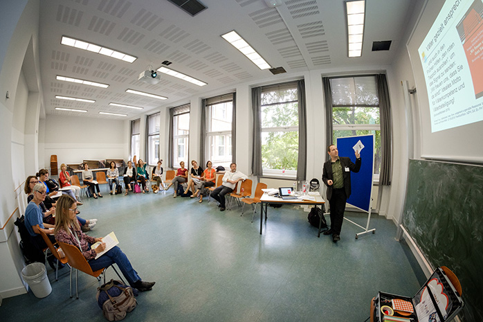 Dr. Michael Tunç steht vor blauer Pinnwand an einem Tisch mit Laptop und deutet mit einem Zettel in der Hand auf eine Projektionstafel, Teilnehmende sitzen in Halbkreis aus Stühlen und blicken zu ihm