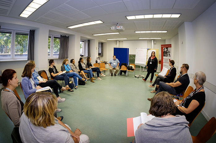 Ruth Römer-Meyer steht vor sitzenden Teilnehmenden in einem Stuhlkreis, hinter ihr stehen Flipcharts und Tafeln