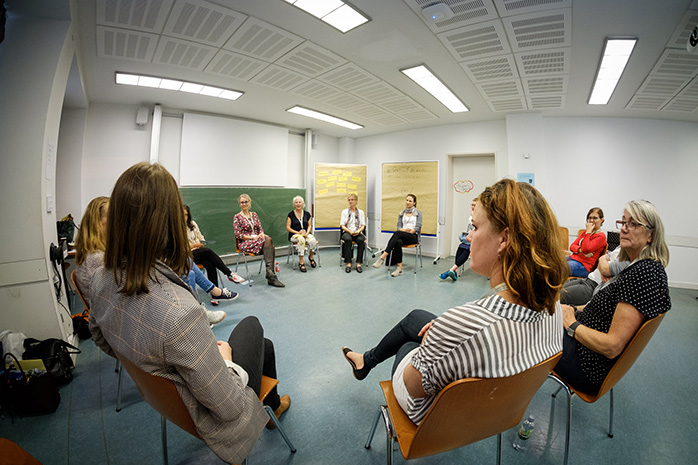 Teilnehmende sitzen in Stühlkreis, im Hintergrund Pinnwände und eine Tafel