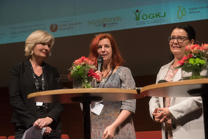 3 Frauen stehen an Stehtischen mit Blumen, eine Frau hält ein Mikrofon in der Hand