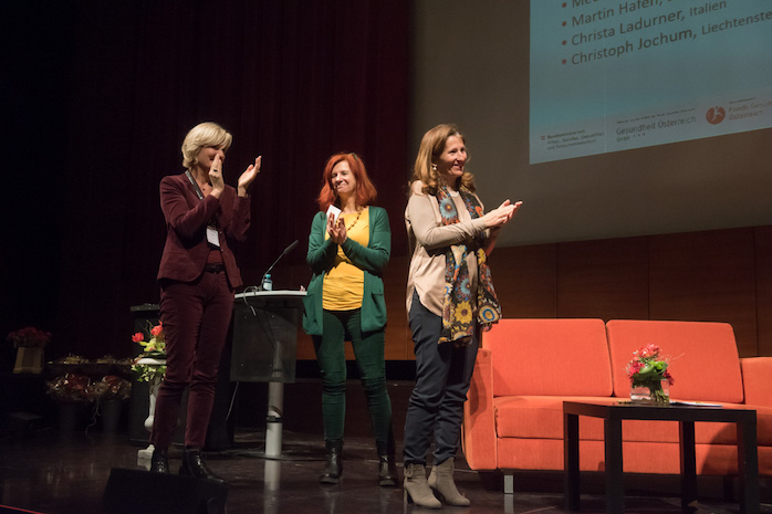 3 Frauen stehen auf der Bühne uns applaudieren, daneben ein rotes Sofa, dahinter eine Präsentationswand