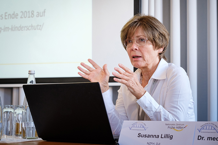 Die Referentin Susanna Lillig sitzt mit Laptop und Namensschild an einem Tisch und gestekuliert mit den Händen, im Hintergrund ein Vorhang mit Projektionswand