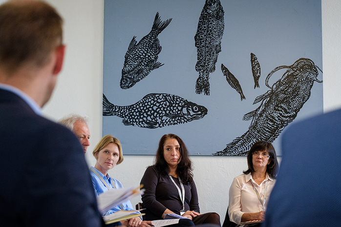 Teilnehmende sitzen zueinander gerichtet, im Hintergrund ein blau-graues Bild mit gemalten schwarzen Fischen