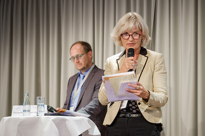 Mechthild Paul steht vor Vorhang mit Mikrofon und Zetteln in der Hand, links hinter ihr sitzt Dr. med. Sönke Siefert an einem Tisch mit weißer Decke und schaut herab