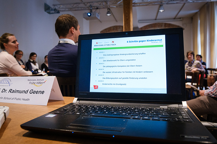 Laptop mit Präsentation auf dem Bildschirm steht auf dem Tisch des Vortragenden, im Hintergrund sitzten Teilnehmende
