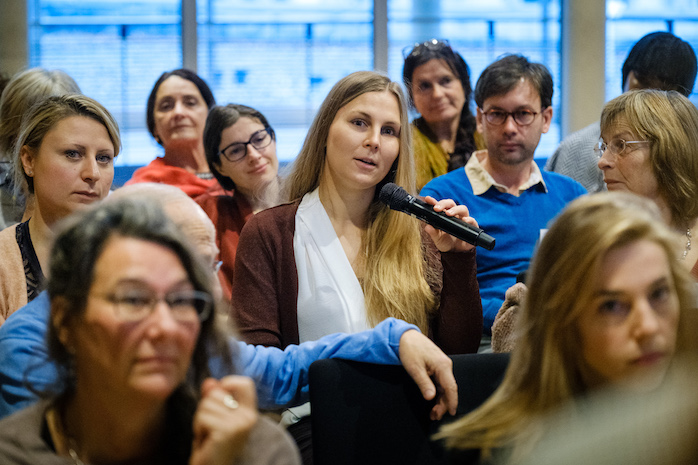 Teilnehmende sitzen auf Stühlen im Plenum, eine Teilnehmerin spricht ins Mikrofon