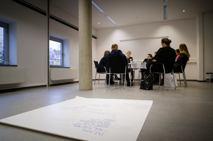 Teilnehmerinnen und Teilnehmer sitzen an einem Tisch, im Vordergrund liegt ein Plakat auf dem Boden