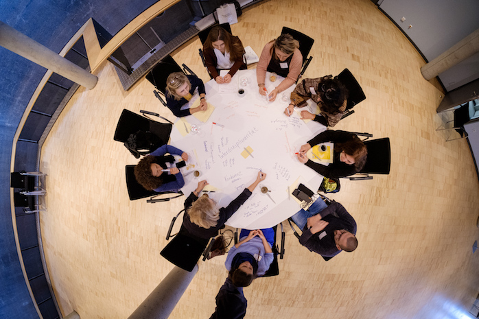 Blick von oben auf einen runden Tisch, um den Teilnehmerinnen und Teilnehmer sitzen